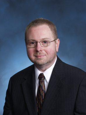 A man wearing a black suit and glasses. A slight smile on his face with a blue background behind him